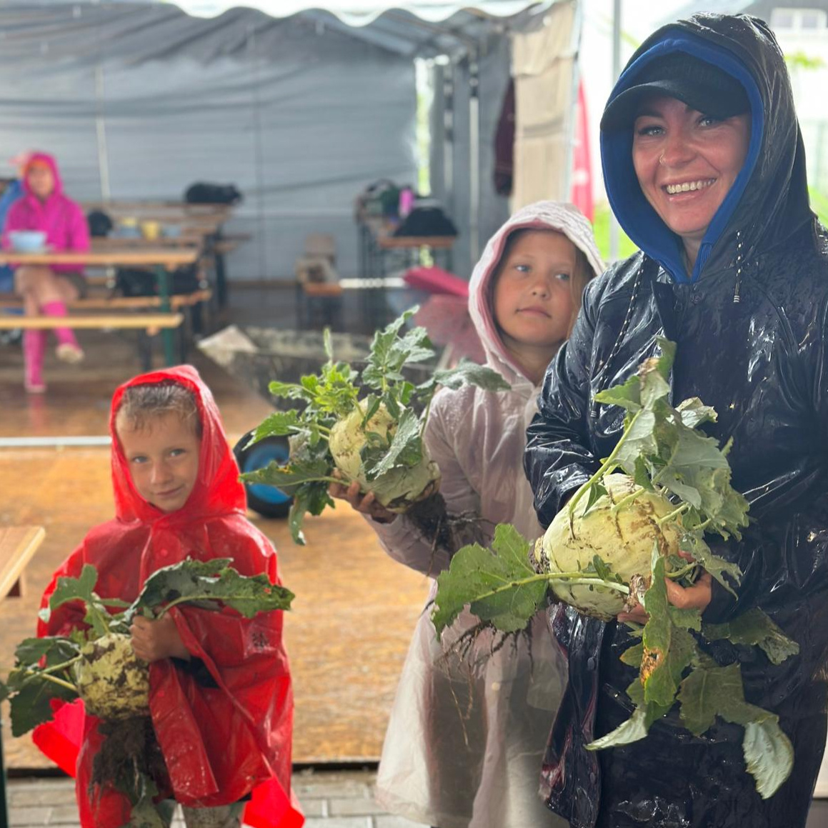 Gartenarbeit bei jedem Wetter. Eine Gruppe von Kindern in Regenmänteln hält zusammen mit einer lächelnden Betreuerin riesige Kohlrabi während einer Aktivität im Freien.