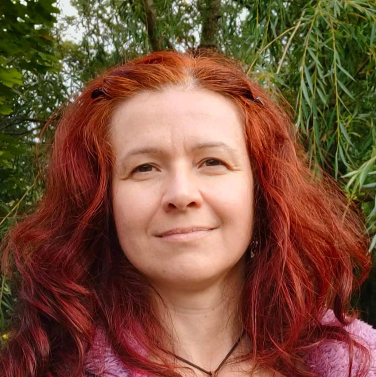 Odzyskaj kontakt ze swoim ciałem. Anna Starba – instruktorka zajęć „Balans Ruch & Relax”, portret uśmiechniętej kobiety o rudych włosach na tle zieleni.